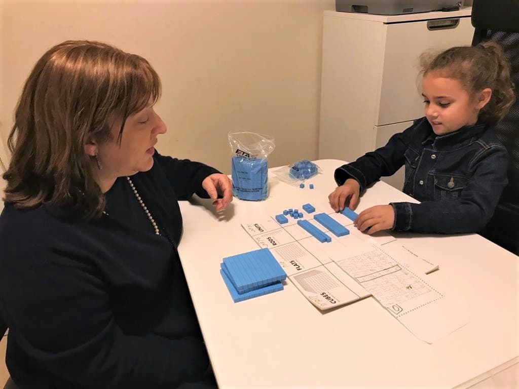 מורה ותלמידה יושבות משני צדי שולחן ומסדרות קוביות בסיס עשר על לוח ערך המקום